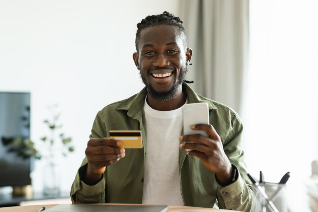 Internet mobile shopping concept. Happy black man using smartphone and holding credit card, purchasing online from home