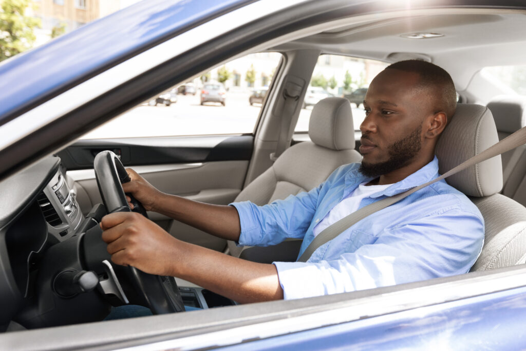 Confident Professional Driver. Side view profile portrait of young African American man sitting in a car on driver's seat. Focused black guy riding in the city, holding hands on steering wheel